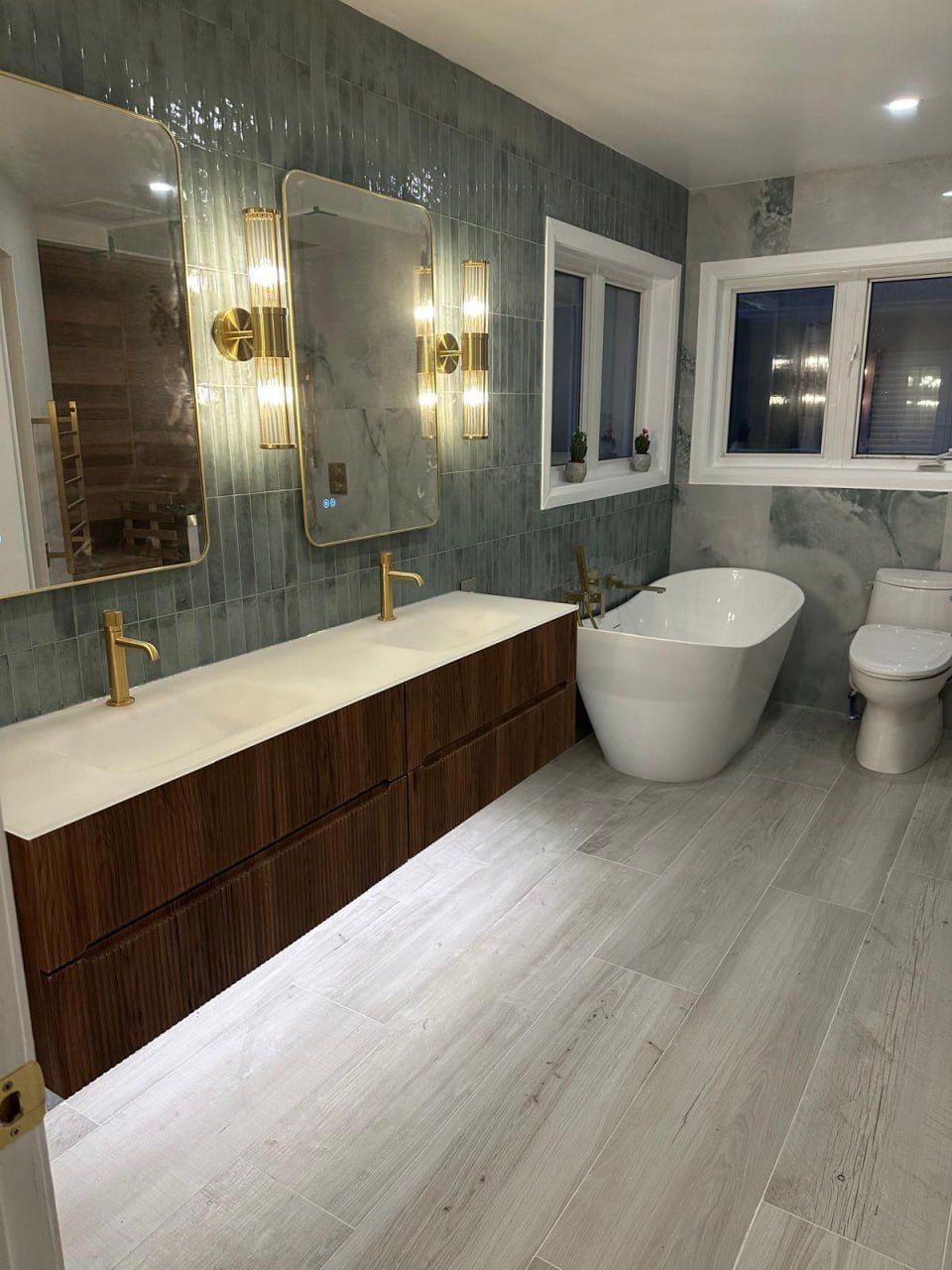 Green-tile ensuite at night with freestanding tub and gold sconces